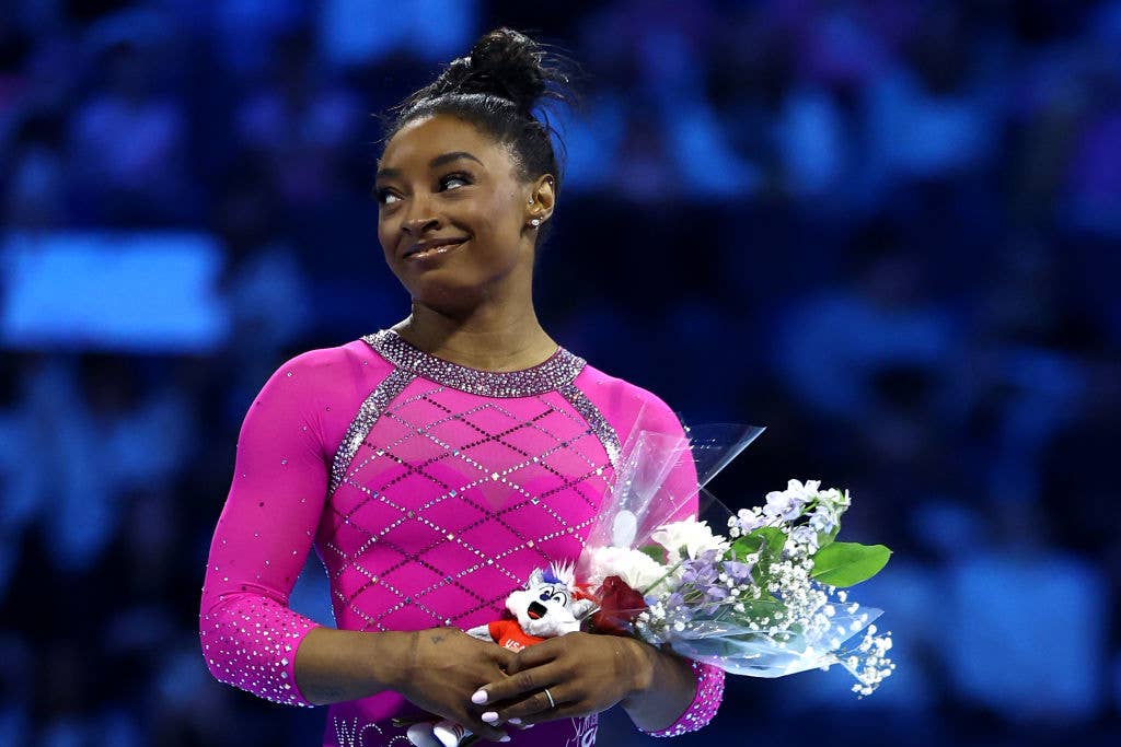 Simone Biles, in a bejeweled leotard, holds a bouquet of flowers and a small plush toy at a gymnastics event. She smiles and looks to the side
