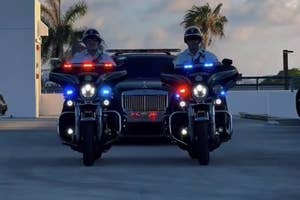 Two motorcycle police officers escort a black luxury car down a sunny, palm-lined street