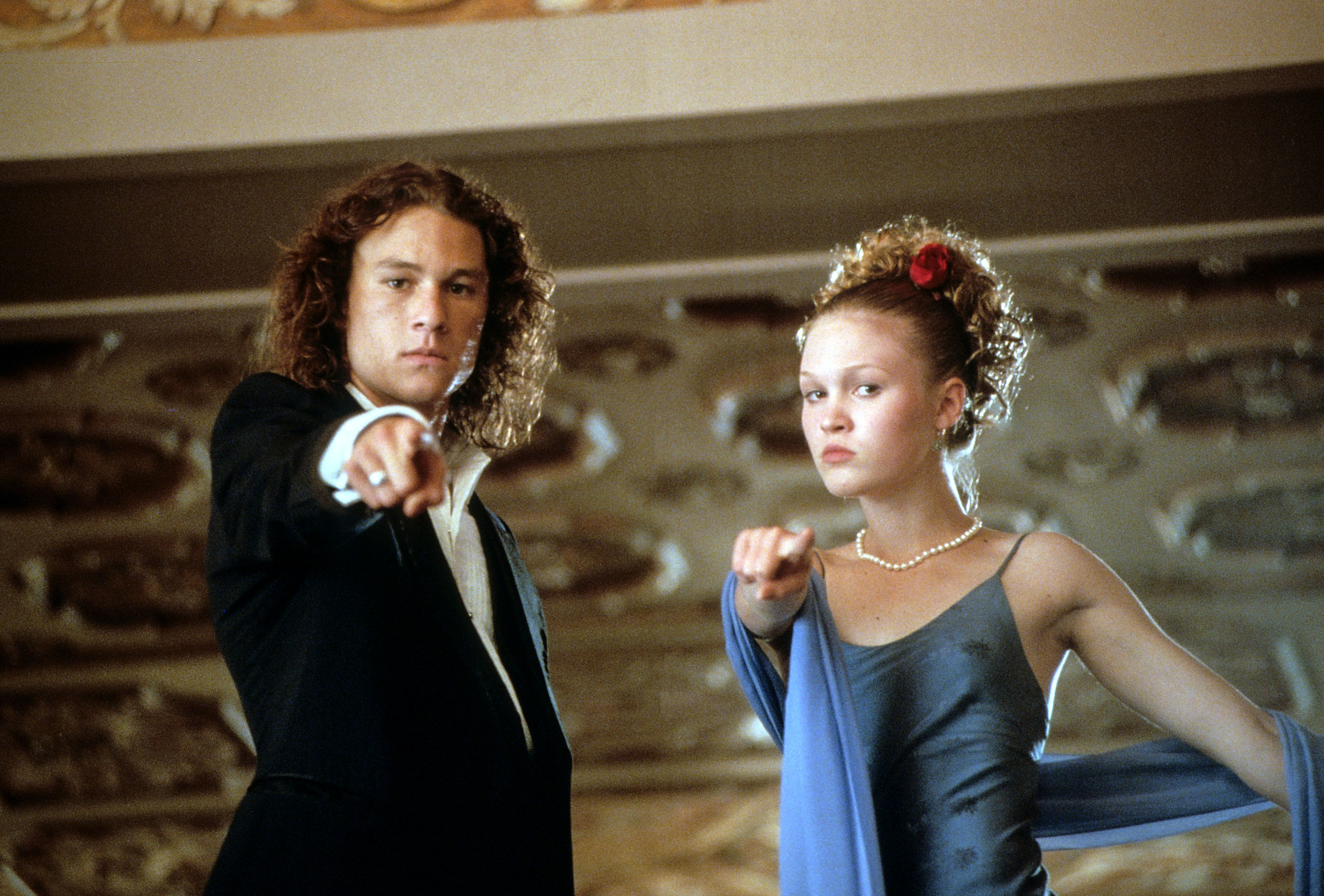 Heath Ledger and Julia Stiles, in formal attire, pointing forward in a scene from a movie. Heath in a suit, Julia in a dress with a shawl