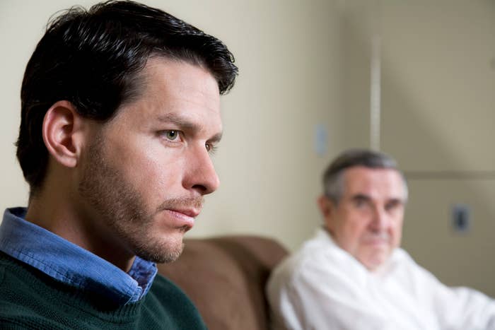 A man in a green shirt and a blue collar appears thoughtful or concerned while an older man in a white shirt looks at him in the background