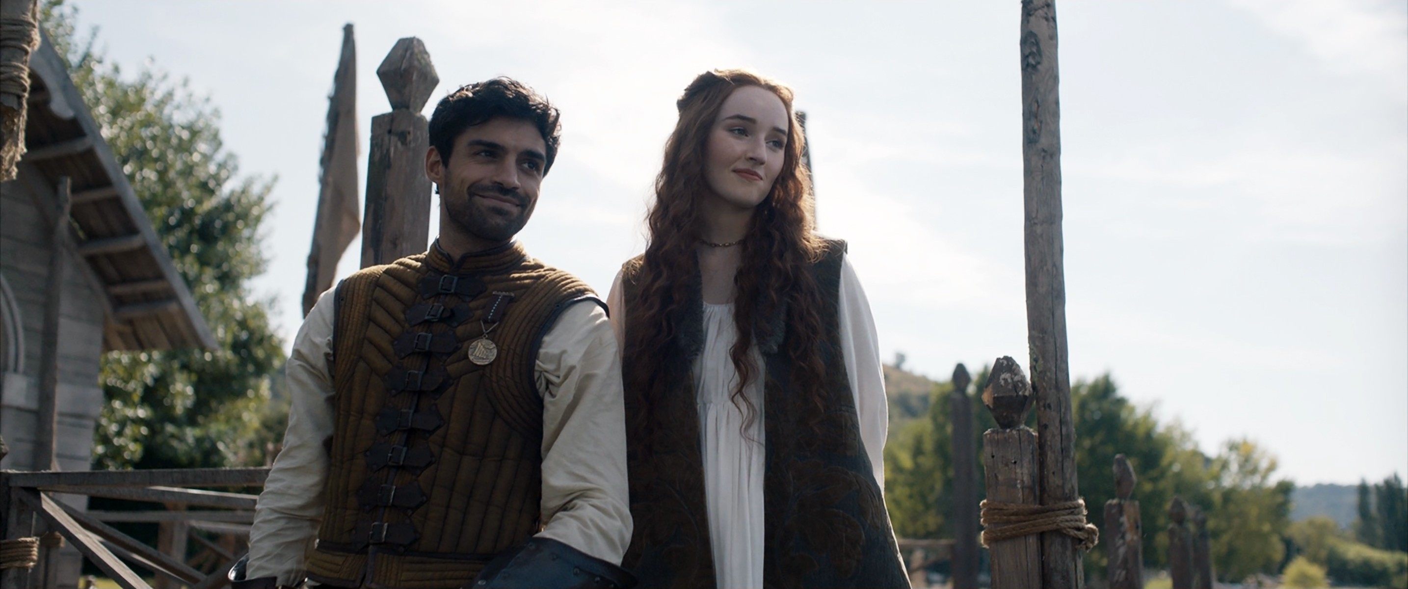 Nida Khurshid and Marisa Devins stand outside, dressed in medieval-style attire with wooden structures and greenery in the background