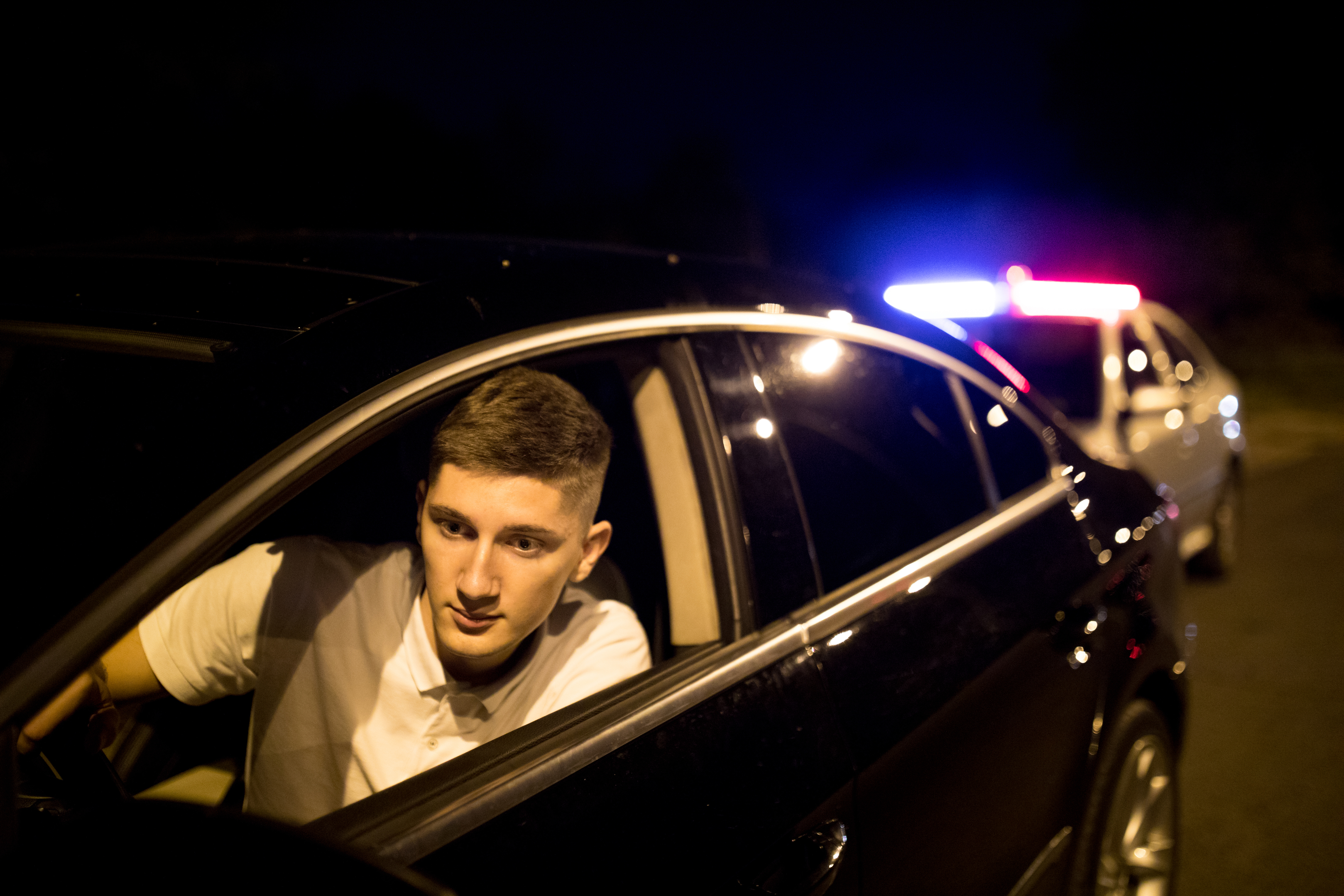 A young man in a white shirt leans out of his car window at nighttime as a police car with flashing lights is stopped behind him