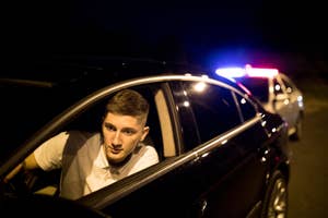 A young man in a white shirt leans out of his car window at nighttime as a police car with flashing lights is stopped behind him