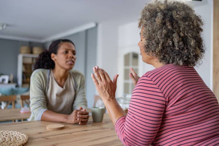 Two women sitting at a table talking in a cozy kitchen. One is gesturing with her hands while the other listens attentively. Names are not identified