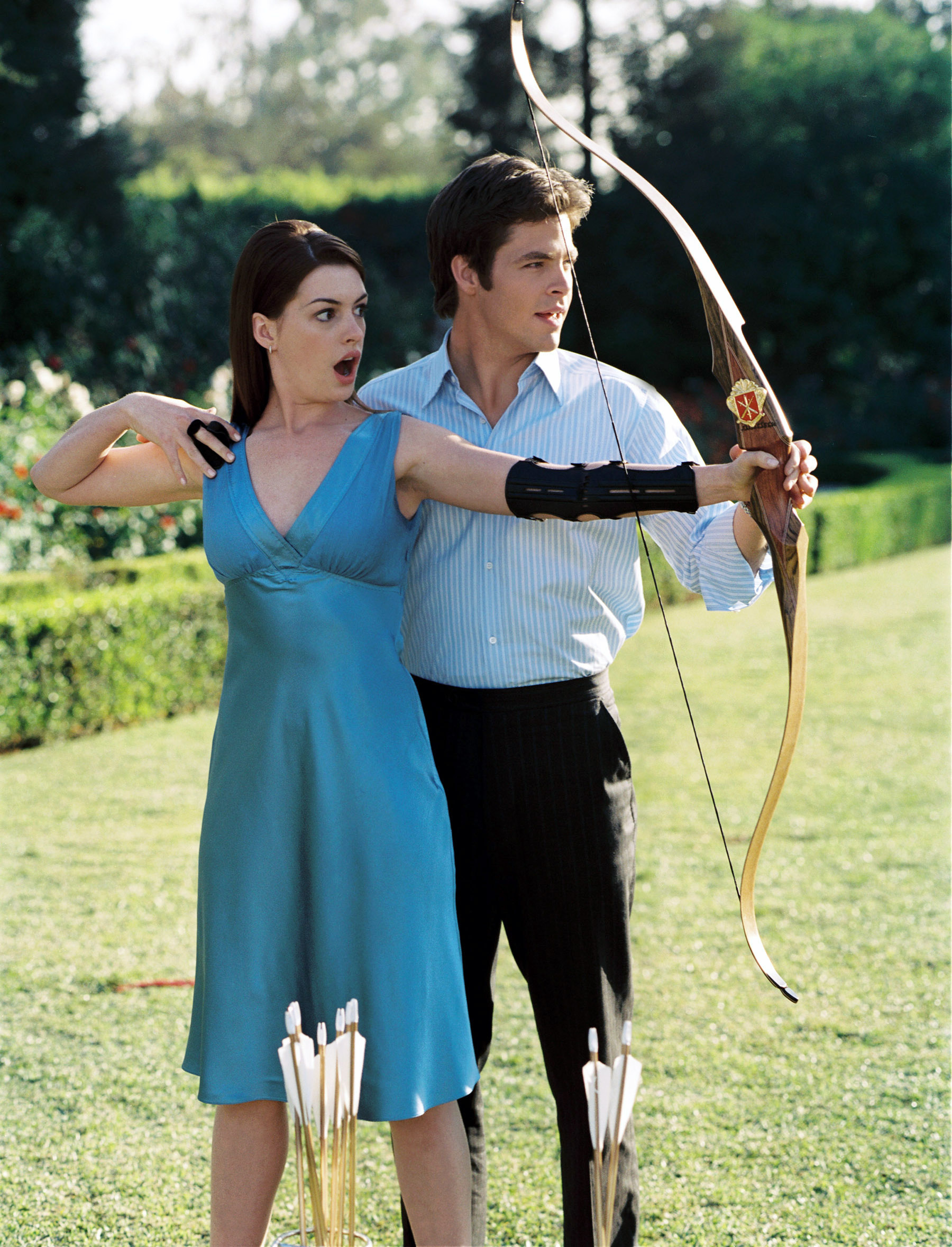 Anne Hathaway helps a man aim a bow and arrow in an outdoor setting, both dressed in casual elegant attire