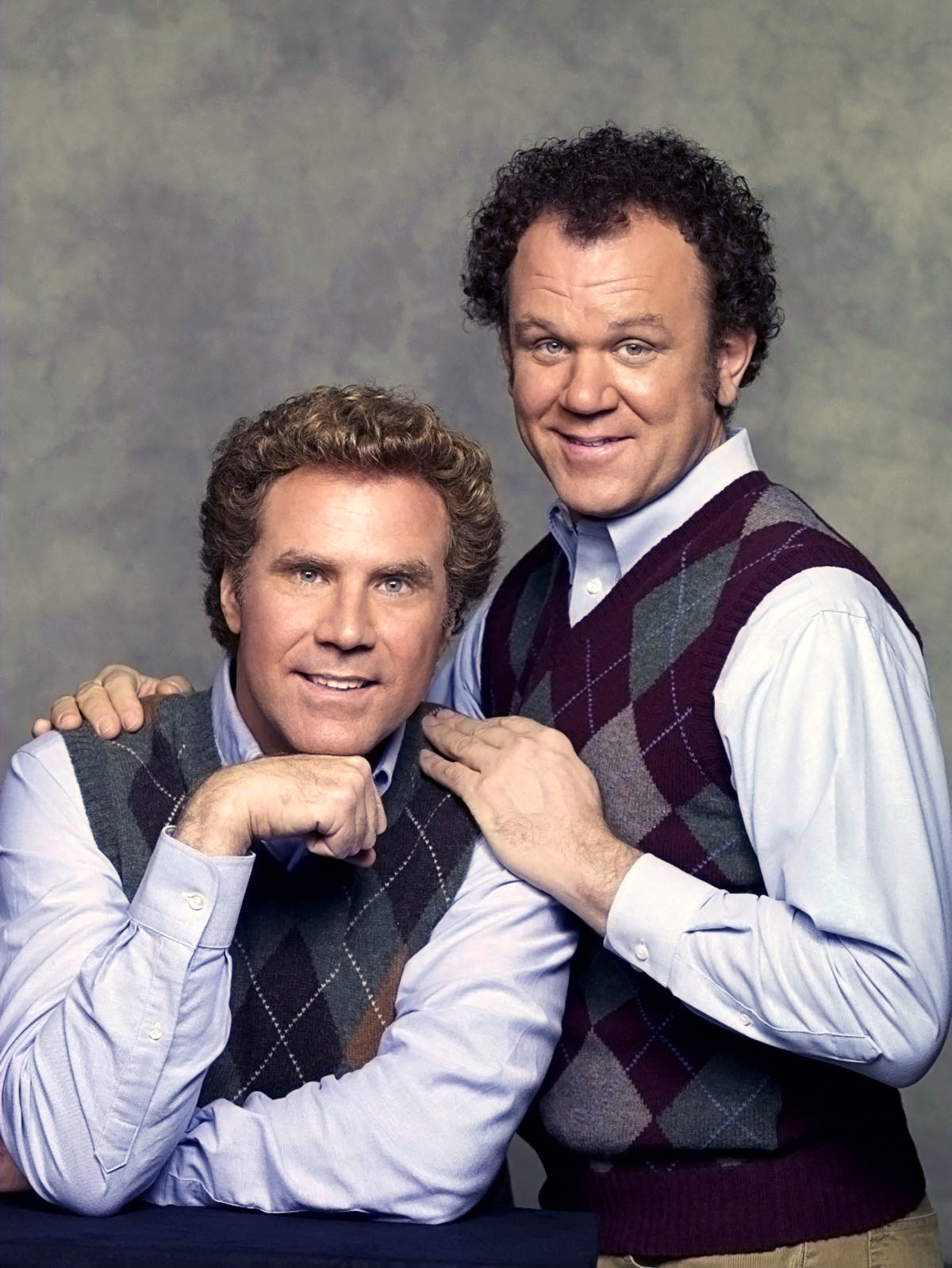 Will Ferrell and John C. Reilly pose together, both wearing light shirts and argyle sweater vests, smiling at the camera against a neutral backdrop