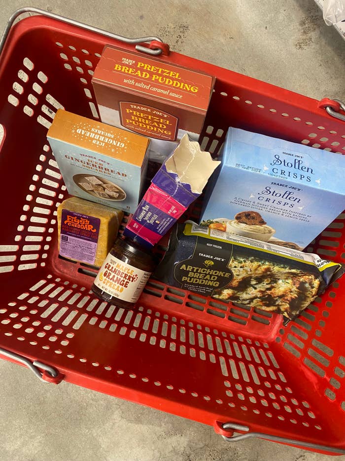 Grocery basket with assorted food items from Trader Joe's including pretzel bread pudding, Stollen crisps, apricot cherry stilton, and artichoke & jalapeño dip