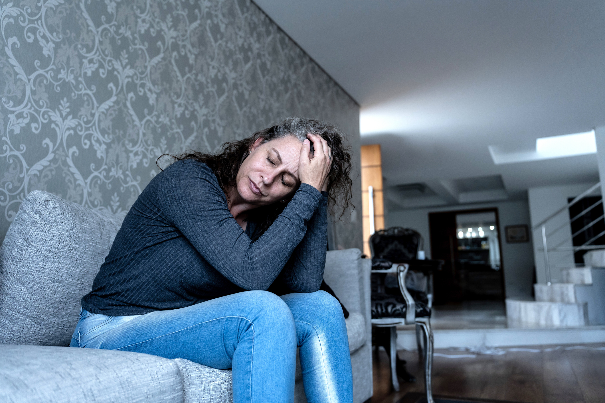 A woman sits on a couch with her head in her hand, appearing distressed or deep in thought, in a modern living room with patterned walls