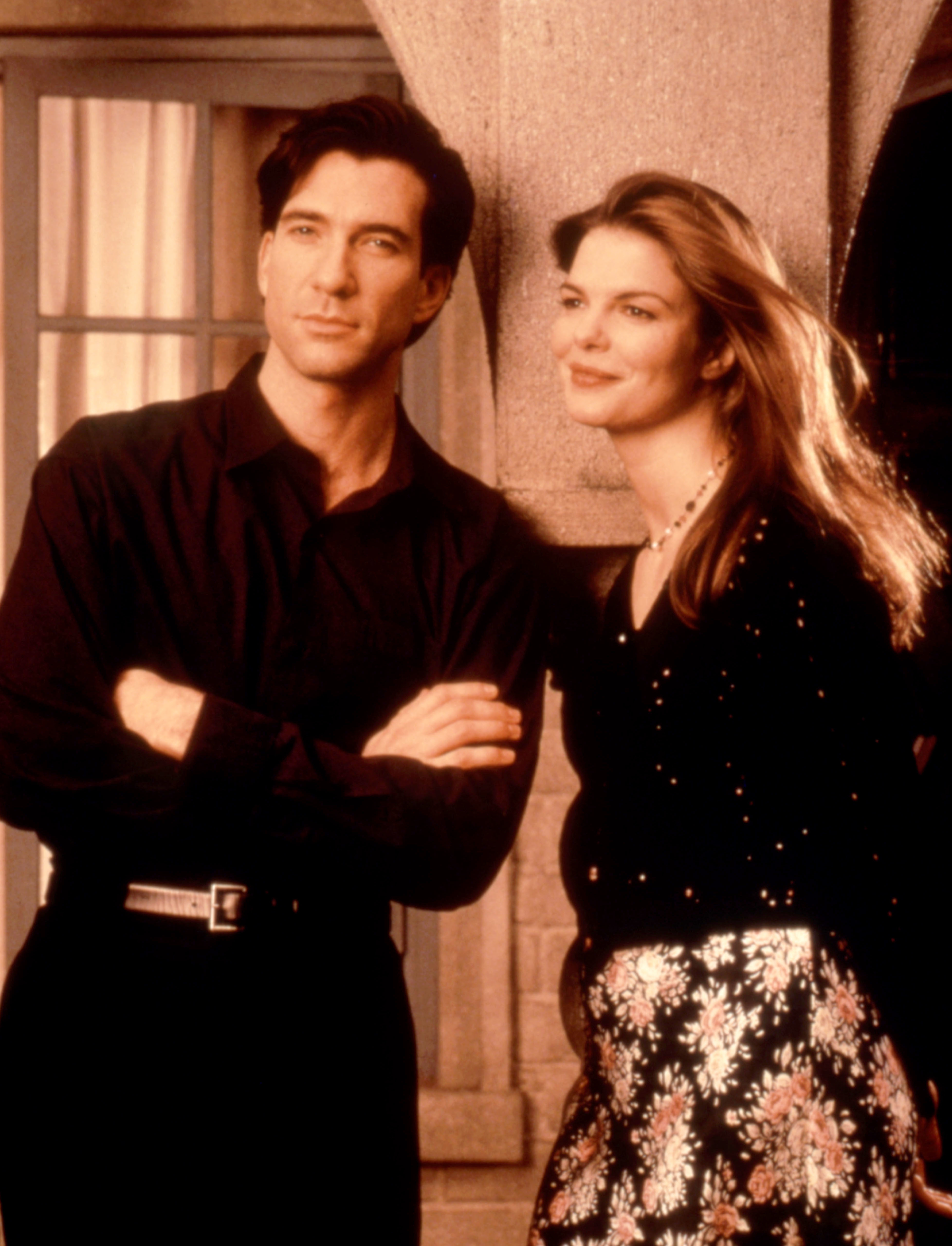 Man and woman posing in front of a column; the man has arms crossed, and the woman has long hair and a floral skirt, both are smiling