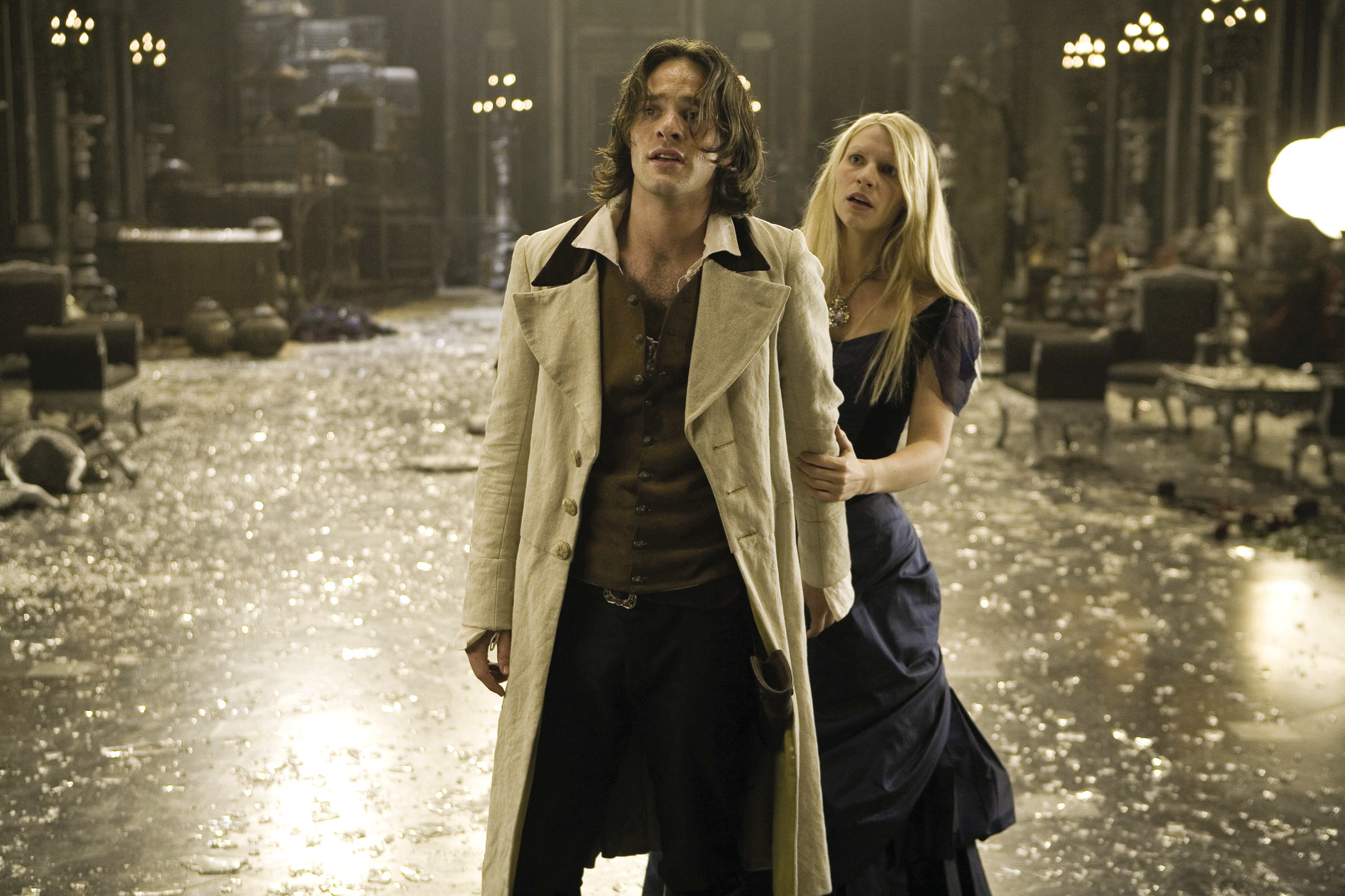 Charlie Cox and Claire Danes in a dramatic scene, with Cox wearing a long coat and Danes holding his arm in a dimly lit room filled with debris