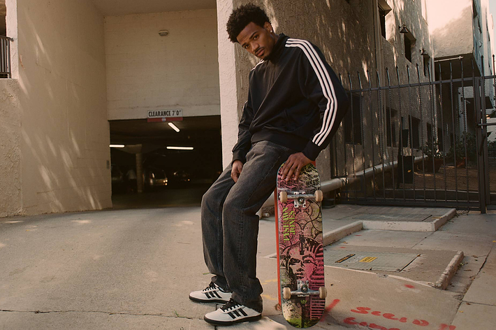 A man stands with a skateboard on the ground, leaning against his leg. He is wearing a black Adidas track jacket, dark jeans, and white Adidas sneakers
