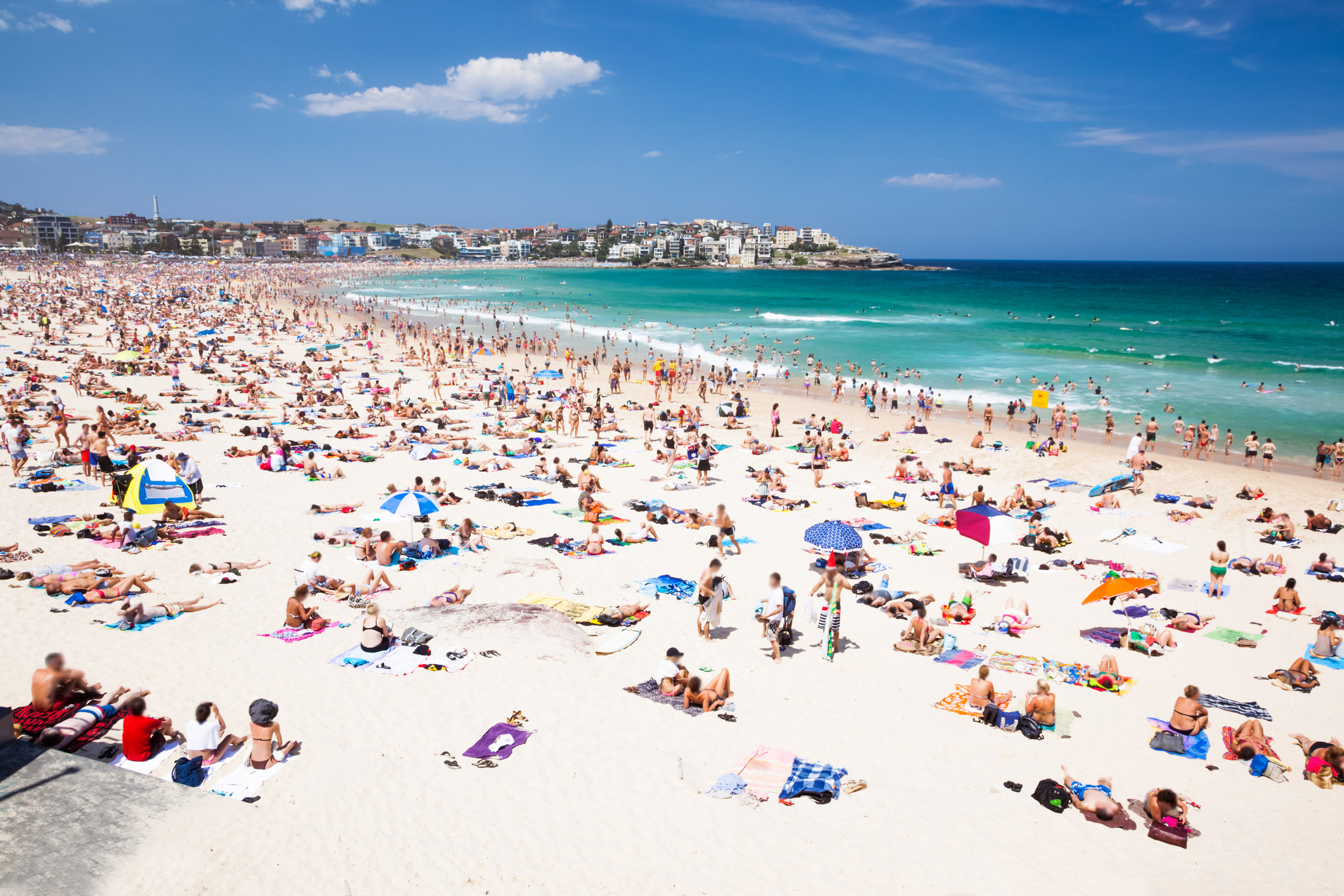 Crowded beach scene with people sunbathing, swimming, and relaxing under umbrellas. In the background, buildings and greenery are visible along the coastline