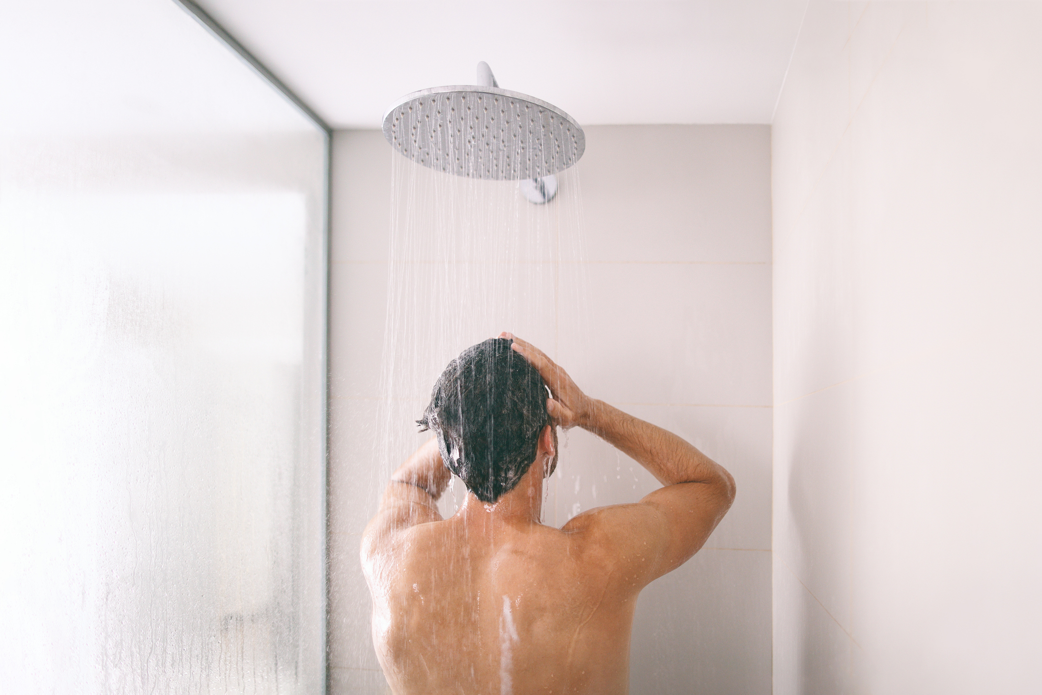 A person showers, facing away from the camera with their hands on their head, under a modern, round showerhead