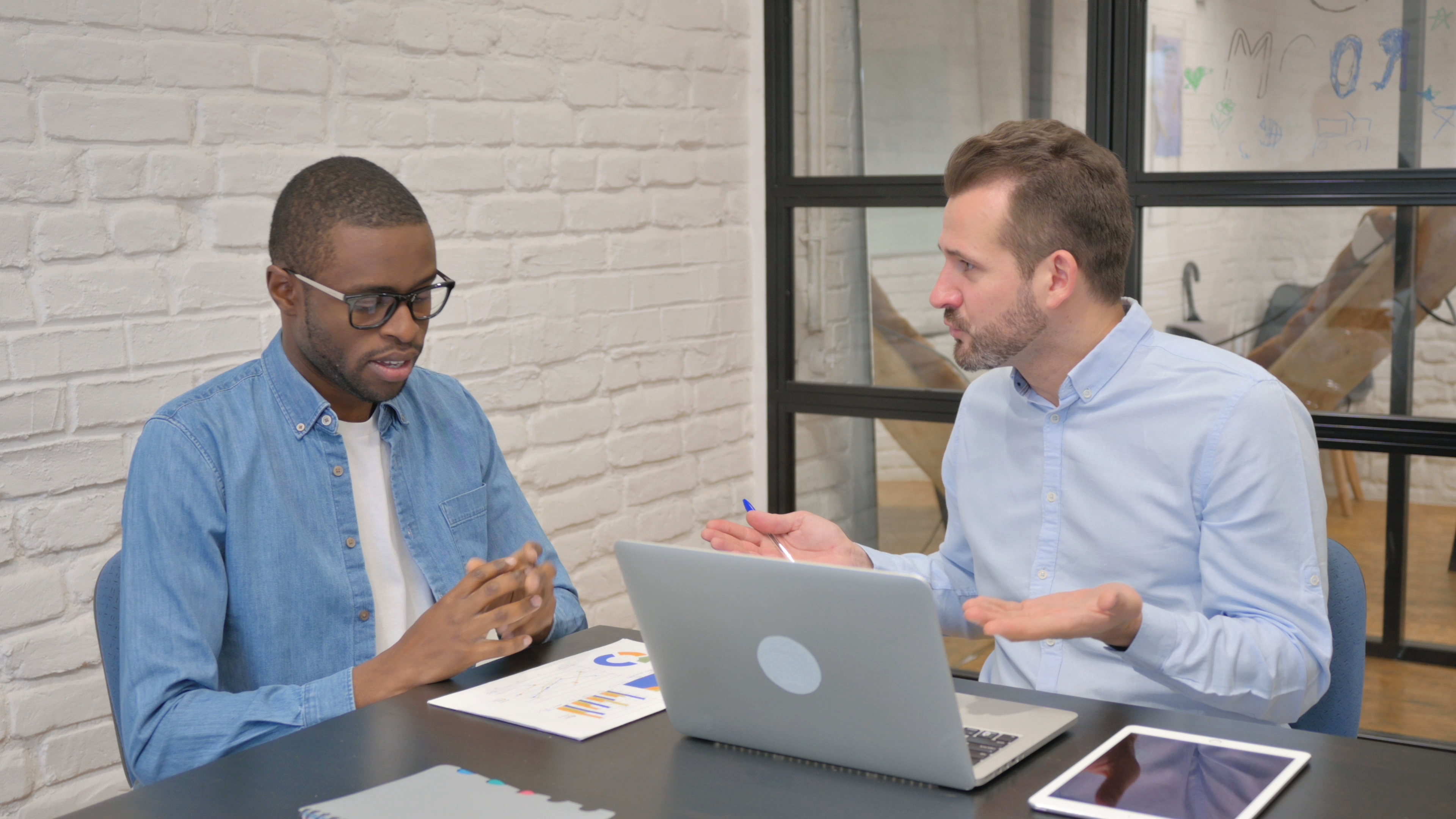 Two men are engaged in a conversation at a desk with a laptop, tablet, and documents. One is presenting while the other listens attentively