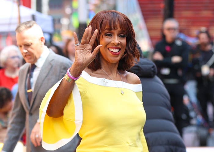 Gayle King waves at a public event, wearing an off-shoulder outfit with flared sleeves. She is smiling, surrounded by people in the background