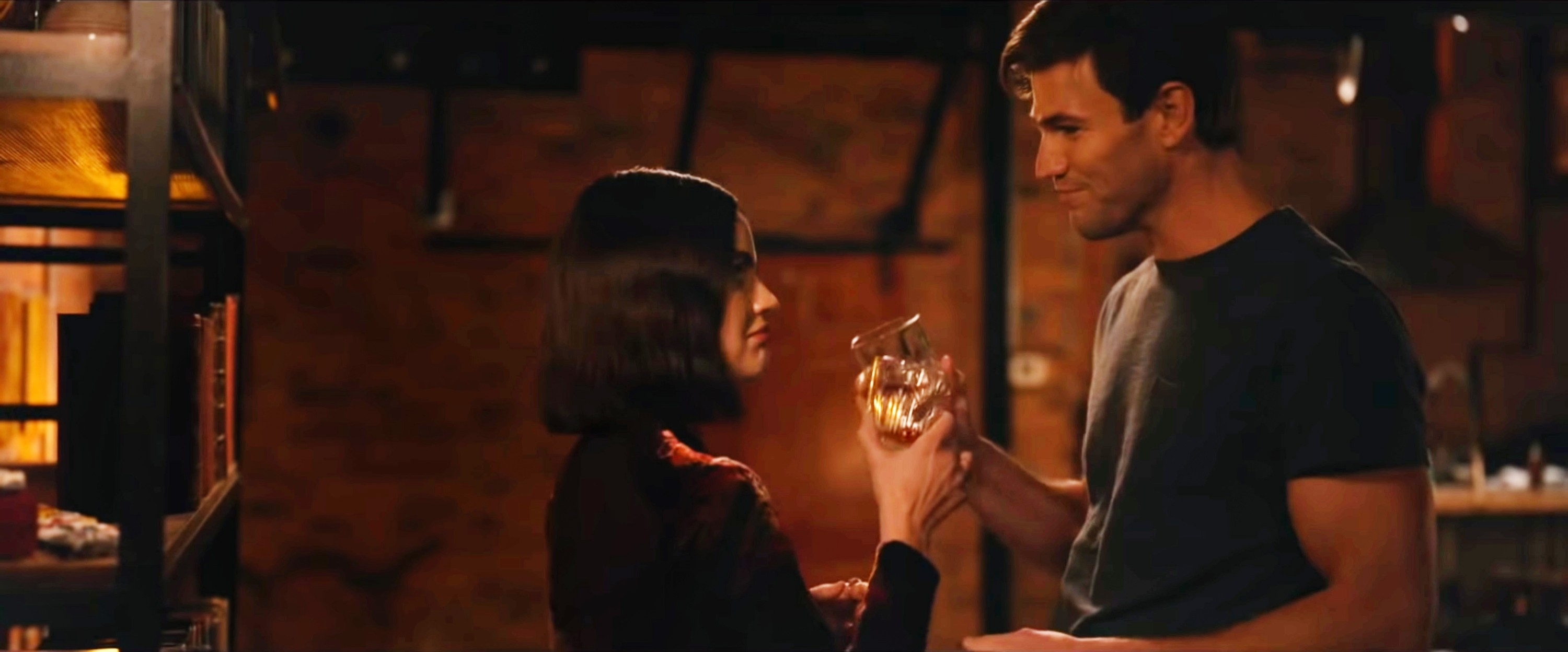 A man and woman, both casually dressed, share a moment while holding a drink in a warmly lit indoor setting