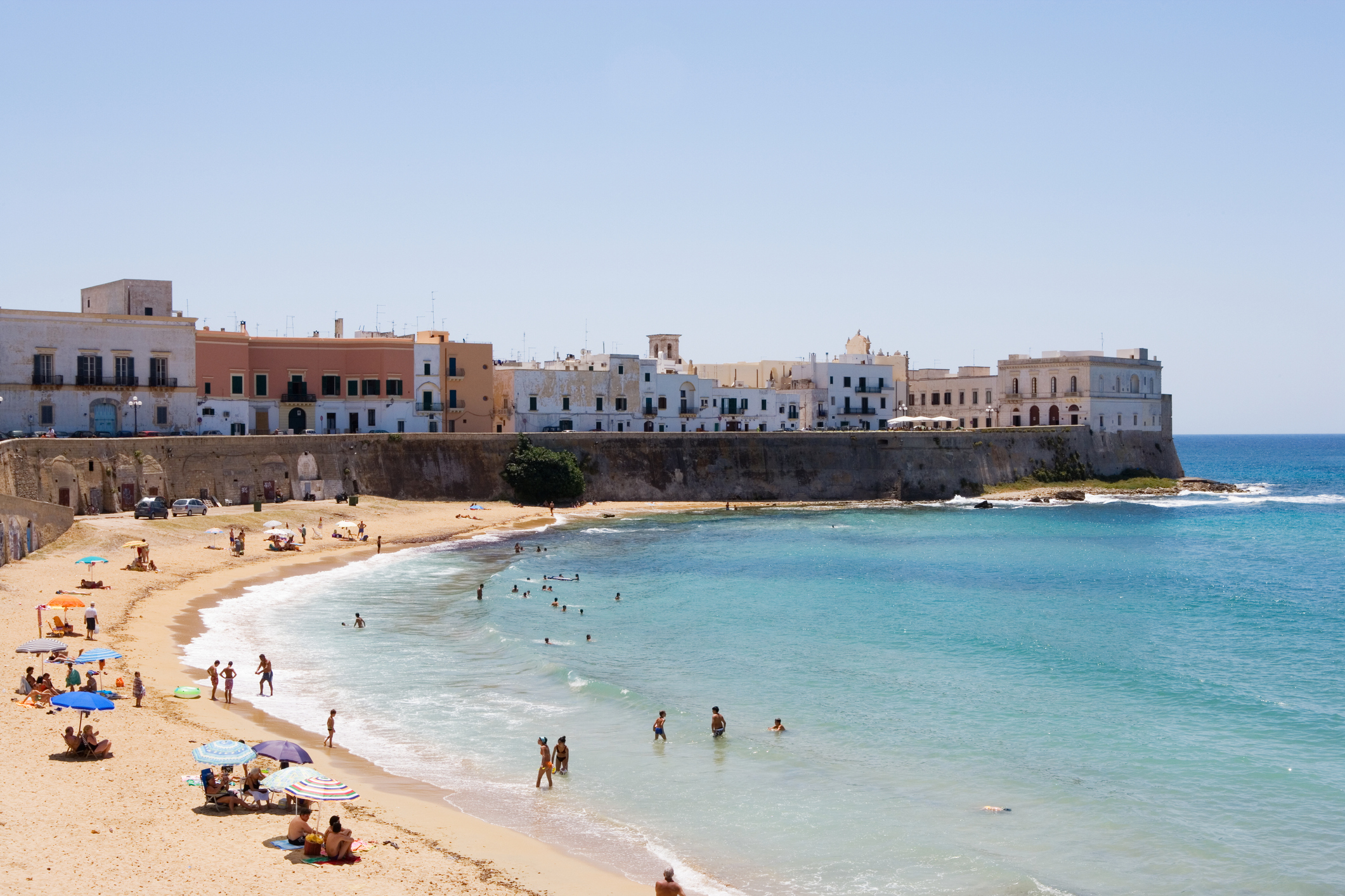 People enjoy a sunny day at a beach with clear water and historic white buildings along the shoreline