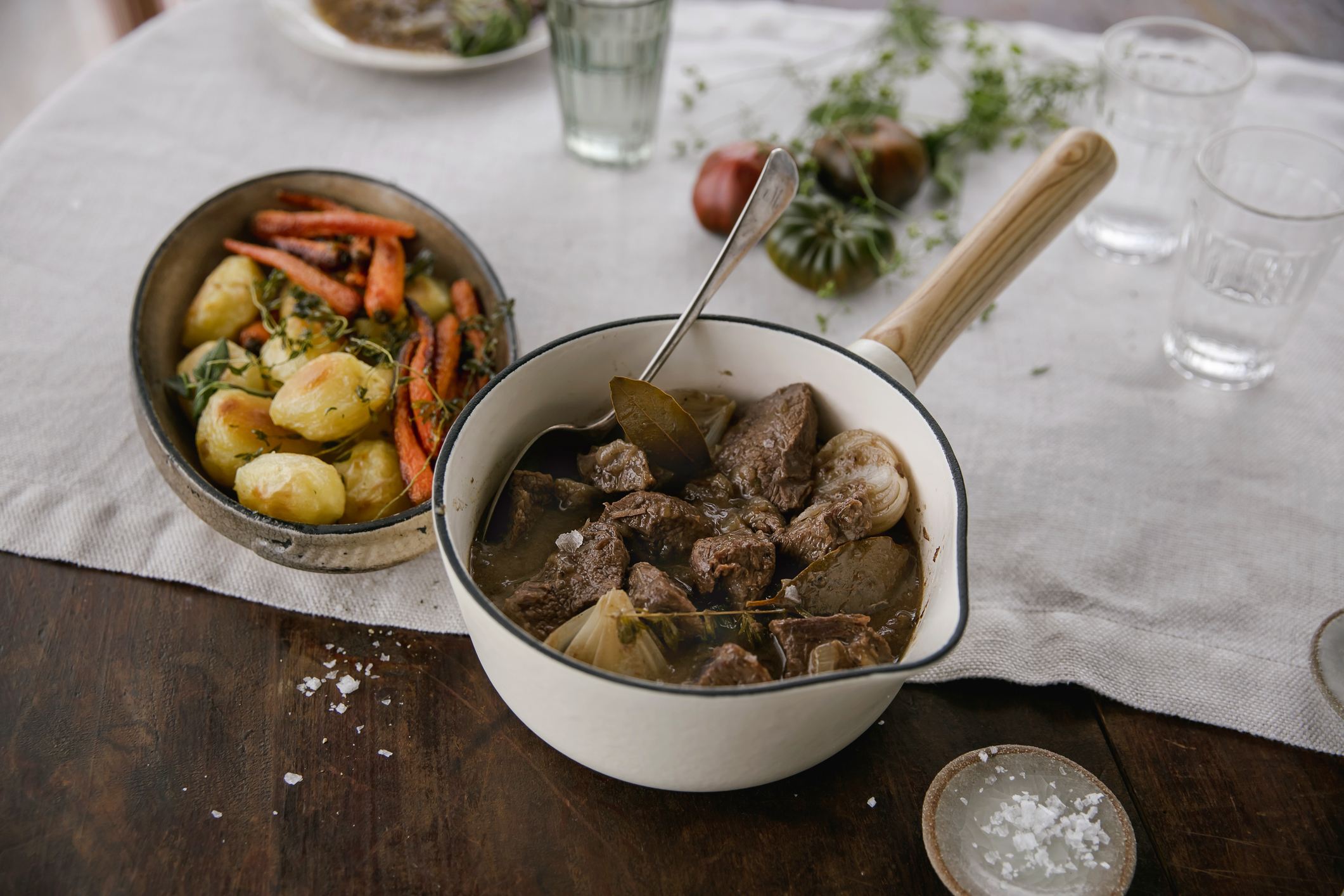 A pot of stew with chunks of meat and onions beside a bowl of roasted potatoes and carrots on a set table