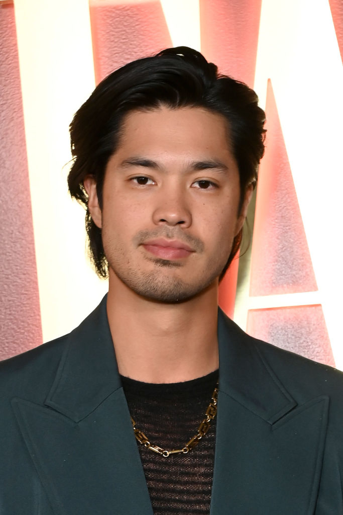 Ross Butler, wearing a dark blazer and a gold chain, smiles at the camera in front of a textured backdrop
