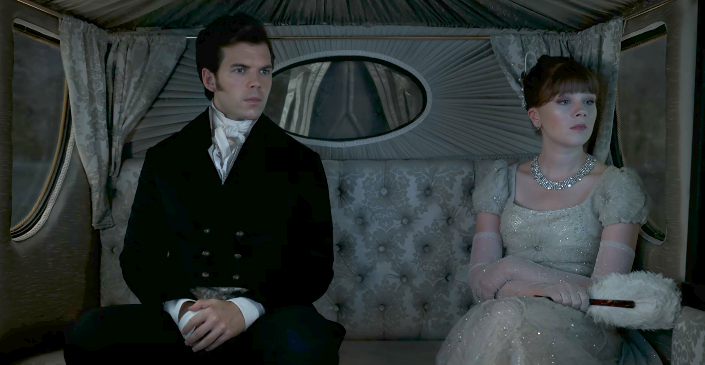 Luke Newton and Claudia Jessie dressed in period clothing, sitting inside a luxurious carriage
