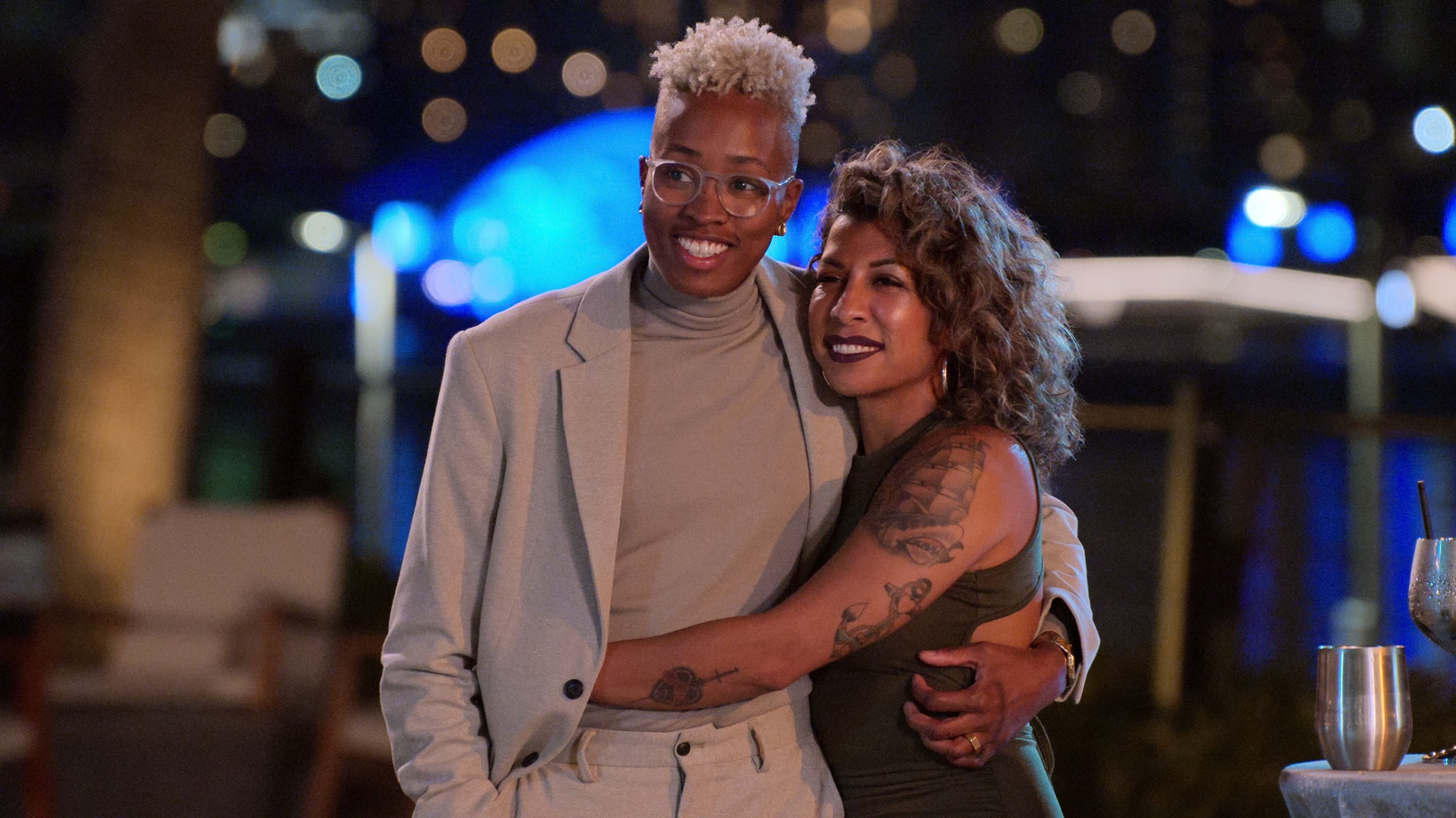 Mal Wright and Yoly Rojas embrace each other, smiling, in stylish evening wear at an event with city lights in the background