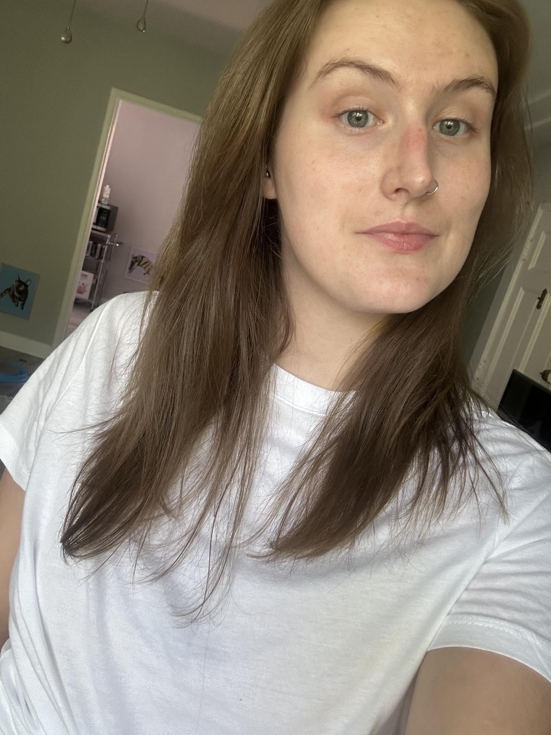A person with long hair, wearing a white T-shirt, looks at the camera in a casual indoor setting