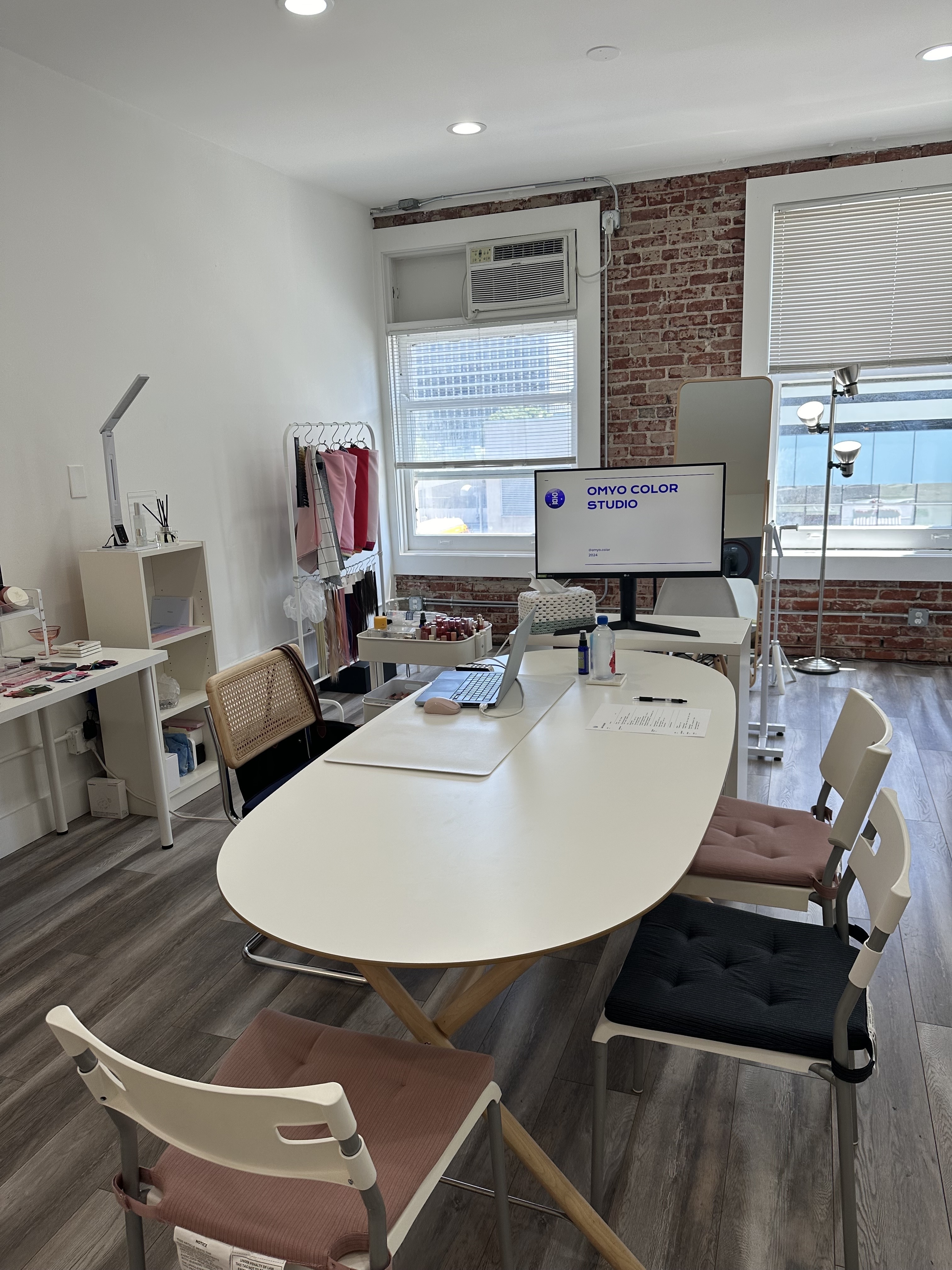 A modern, well-lit office with a white table, chairs, computer monitor displaying &quot;ONYO COLOR STUDIO,&quot; various office supplies, and a clothes rack in the background