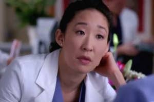 Sandra Oh dressed as a doctor in a scene from Grey's Anatomy, sitting at a desk and looking thoughtfully at someone out of the frame