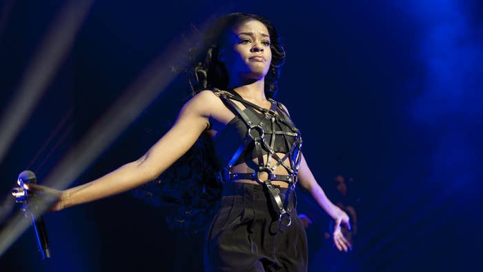 Azealia Banks performing on stage, wearing a black leather harness-style top and black pants, holding a microphone