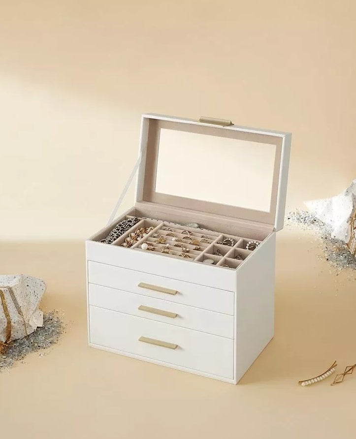 White jewelry organizer box with three drawers, holding various jewelry pieces including bracelets, rings, and earrings; stones and crystals placed around it