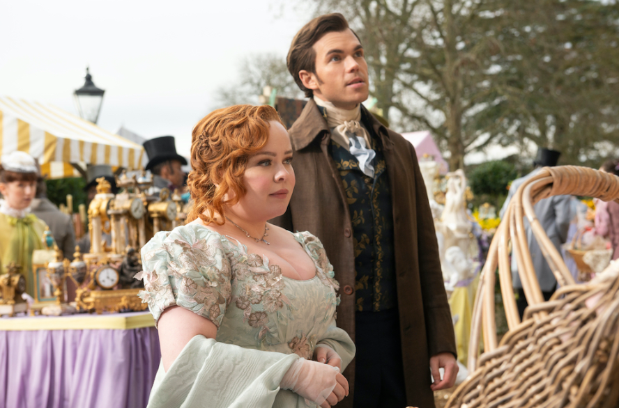 Nicola Coughlan y Luke Newton en trajes de época en un mercado al aire libre lleno de antigüedades. Escena de la serie 'Bridgerton'