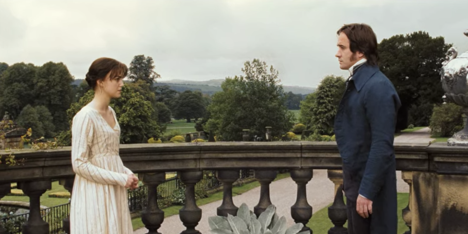 Keira Knightley y Matthew Macfadyen en una escena de película de época, conversan en un balcón con un paisaje de jardín al fondo