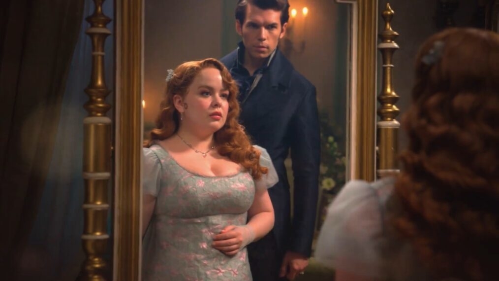 Nicola Coughlan y Luke Newton en una escena de un drama de época, con Nicola mirando hacia un espejo grande y Luke de pie detrás de ella