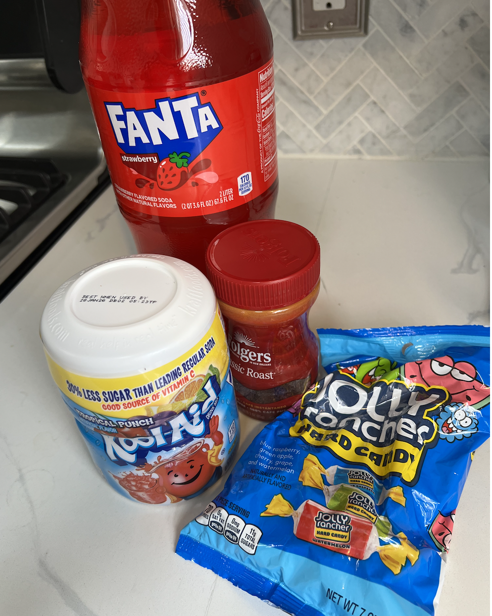 As sealed 2-liter bottle of Fanta strawberry soda, Folgers Classic Roast instant coffee, Kool-Aid mix, and Jolly Rancher hard candy on a kitchen counter