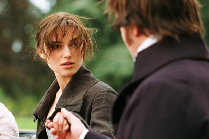Keira Knightley, con peinado desordenado, en una escena de una película basada en una novela. Ella mira a un hombre de espaldas que sostiene su mano