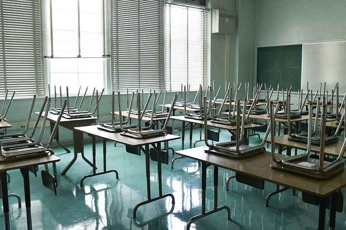 Desks with chairs stacked upside down in an empty classroom with sunlight coming through the window blinds