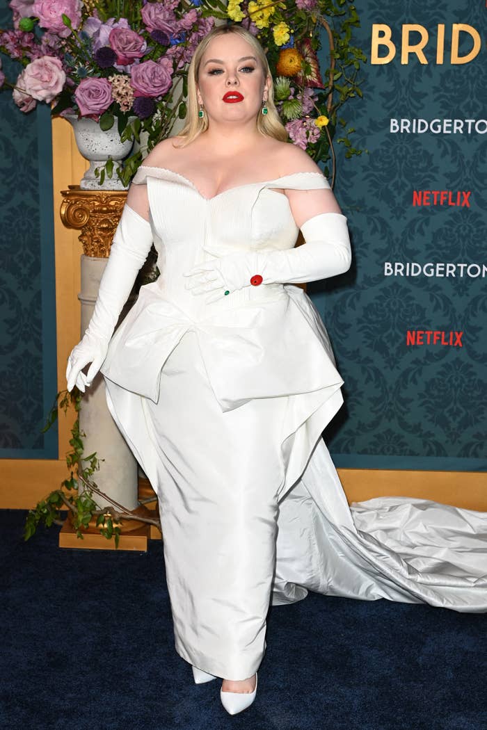 Nicola Coughlan poses on the red carpet in an off-shoulder, white gown with a train, gloves, and matching heels at a Netflix event for Bridgerton
