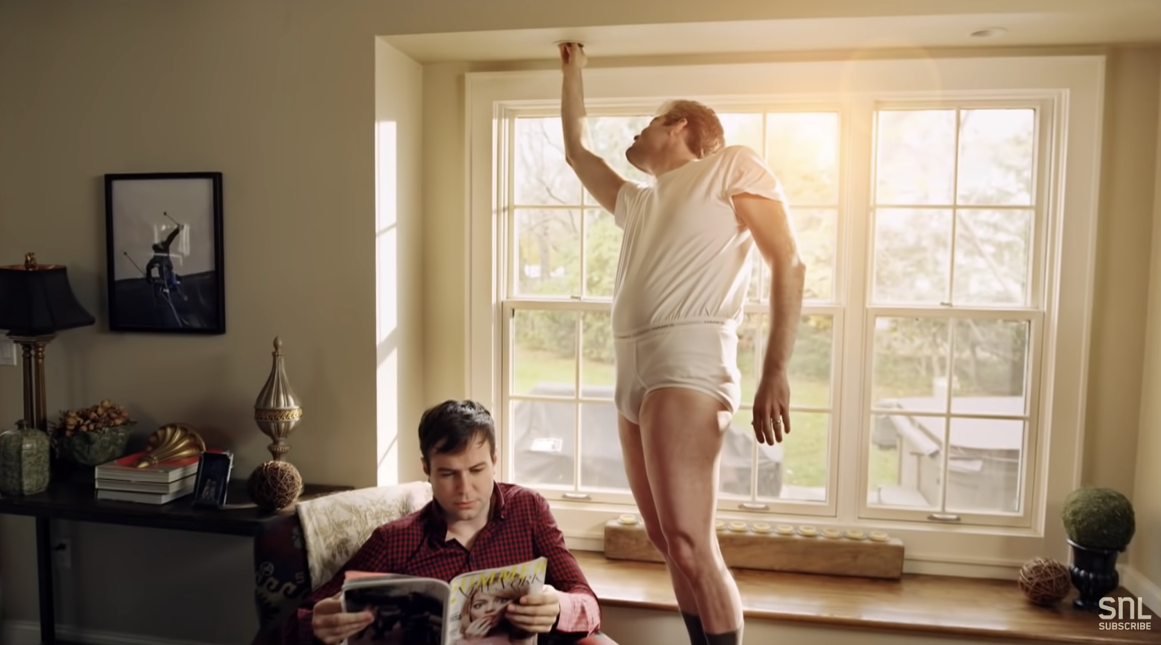 An SNL sketch: A man reads a magazine on a sofa while another man, wearing only white undergarments, stands on a windowsill and reaches for the top of the window