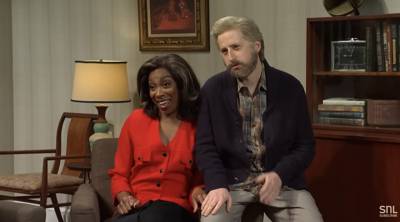 Ego Nwodim and James Austin Johnson in a living room setting, sitting close on a couch during an SNL sketch. Ego is in a red blouse, and James has a gray hair wig with a cardigan