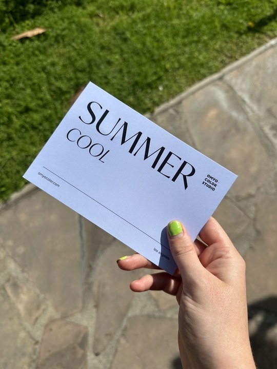 A hand with green nail polish holds a card reading &quot;SUMMER COOL, OMYO COLOR STUDIO&quot; on an outdoor stone pavement next to grass