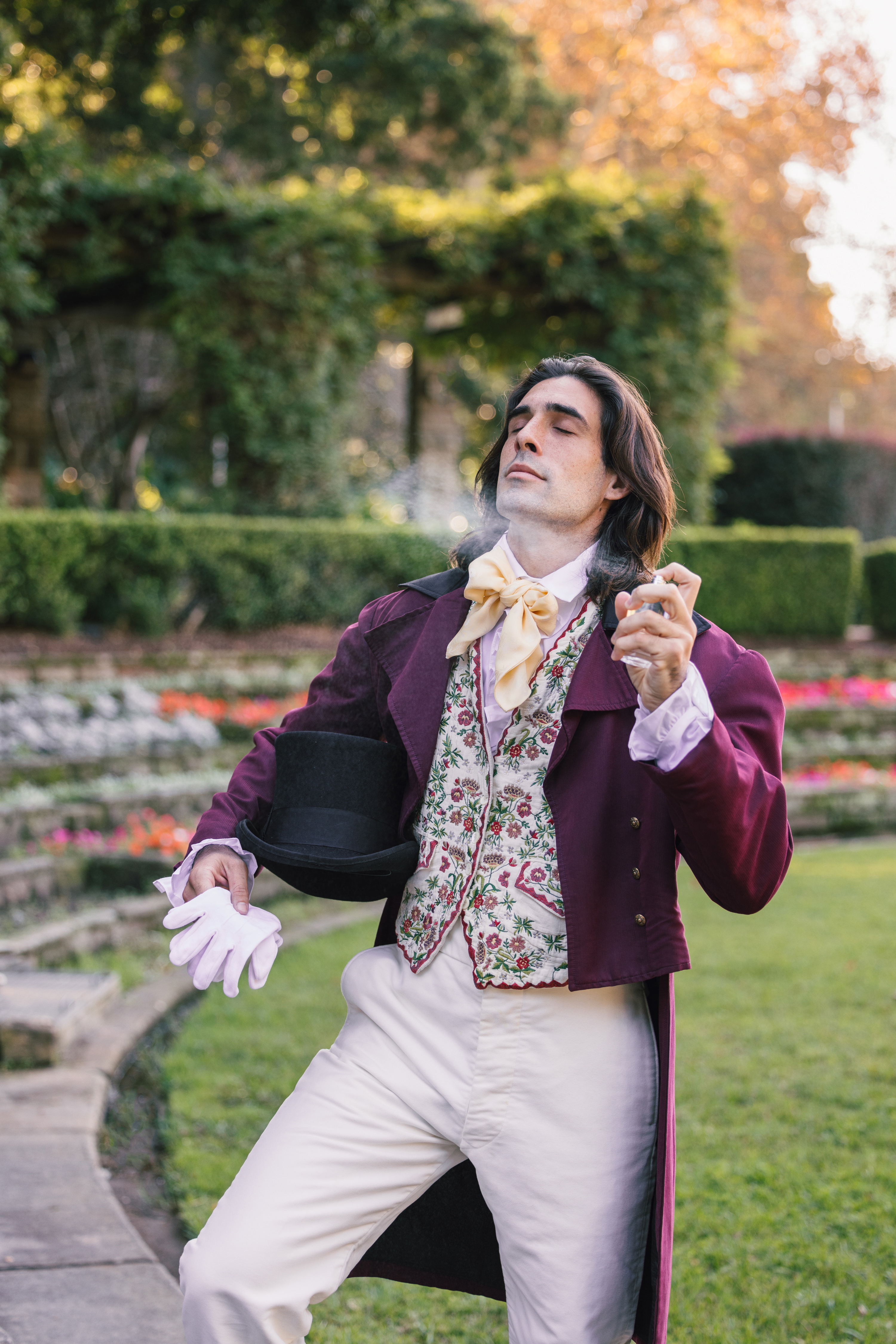 Man dressed in Victorian-style attire with a floral vest and tailcoat, holding a top hat in a garden setting