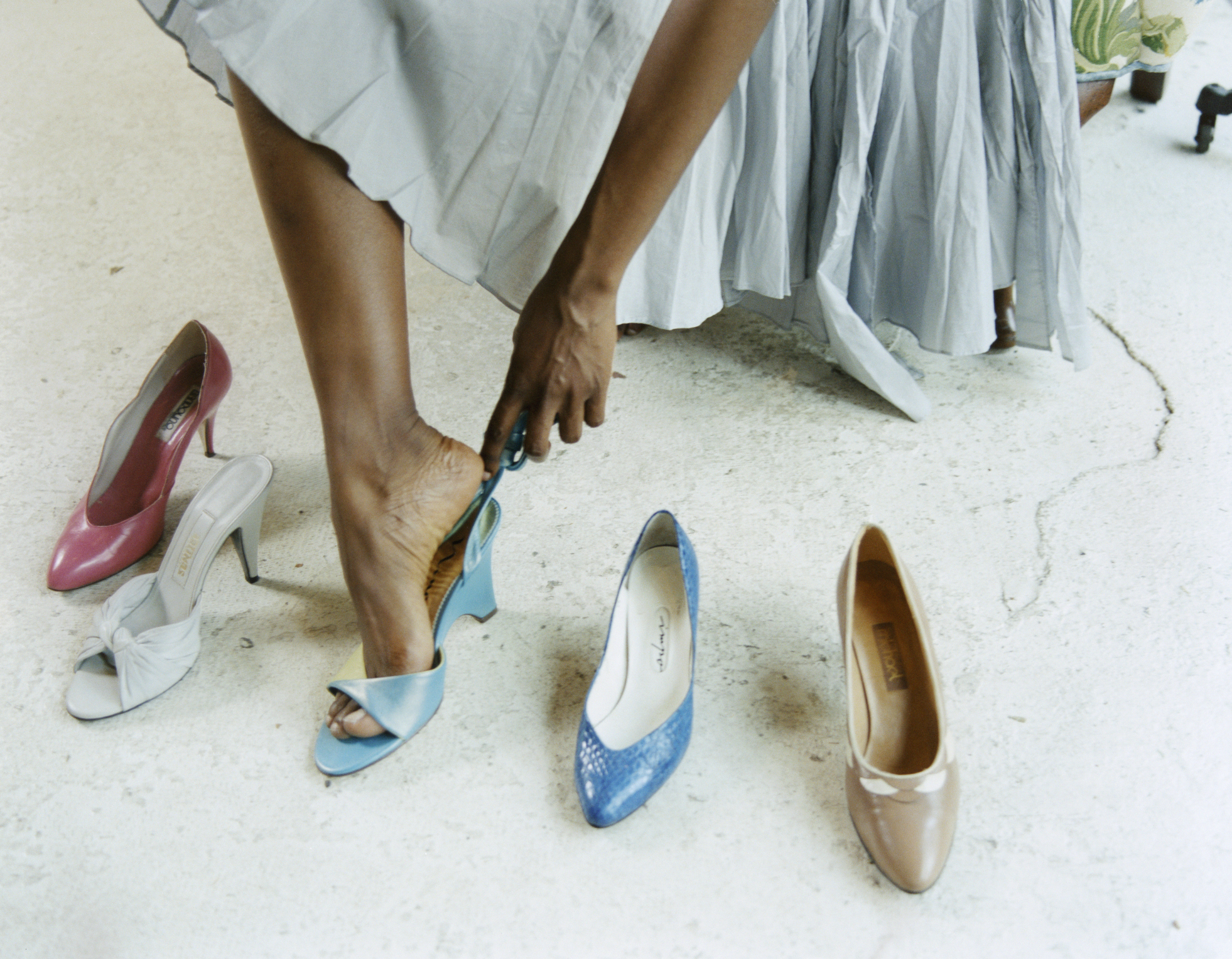 A person trying on different high-heeled shoes including blue, pink, white, and beige. The person is wearing a flowing skirt