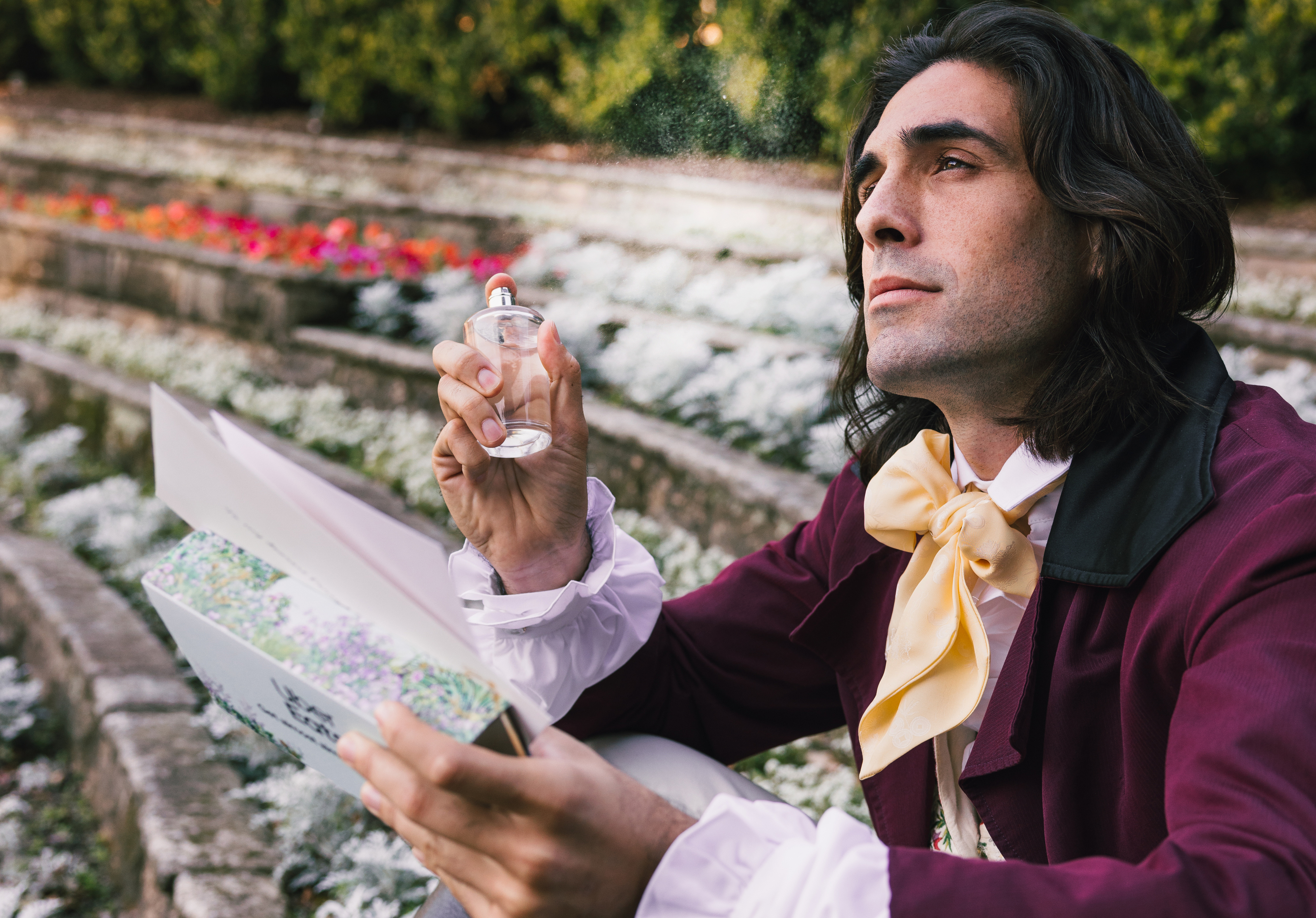 A person in historical attire with a large yellow bow tie holds a perfume bottle in one hand and a book in the other, looking thoughtfully into the distance