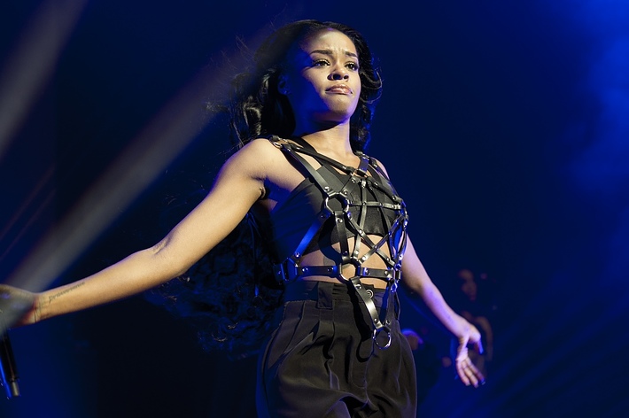Azealia Banks performs on stage wearing a sleeveless top with a strappy harness and high-waisted pants. She has long hair flowing behind her