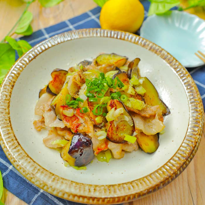 レモンと青ネギを添えた鶏肉と茄子の料理が白い皿に盛られています。背景にナプキンと皿があります。