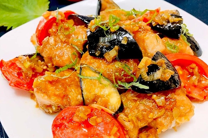 白い皿に盛られた焼き魚とトマトの料理。トマト、なす、ネギが飾り付けられている。