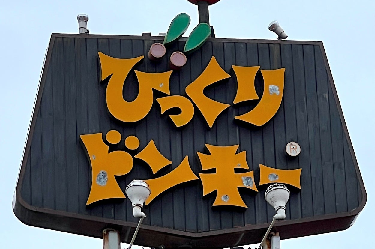 Sign with yellow Japanese characters on a dark background, featuring a red and green stylized figure at the top