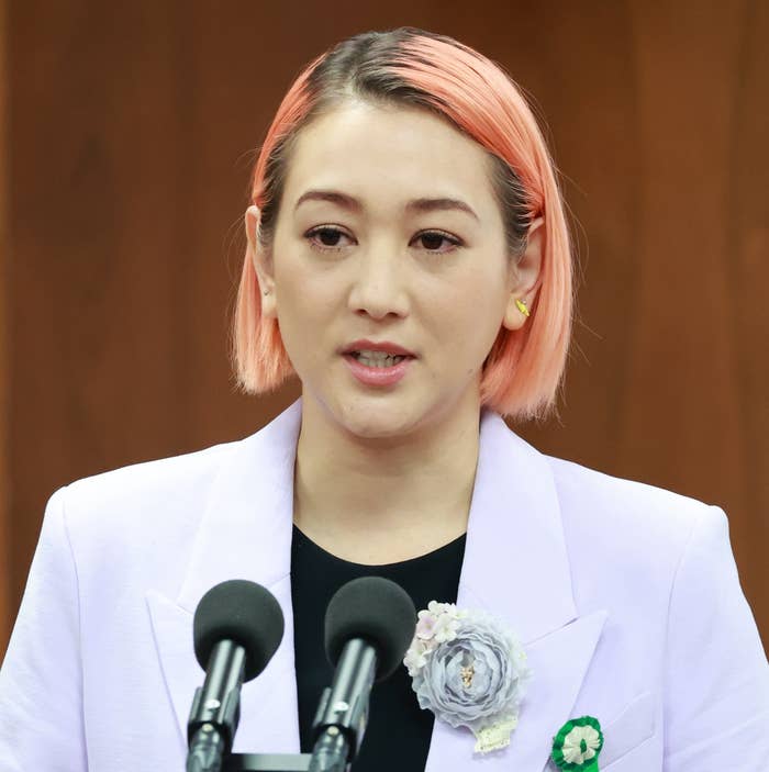 ピンクのショートヘアの女性、川崎希さんがスピーチを行っています。薄紫色のジャケットを着用し、2つのマイクの前に立っています。