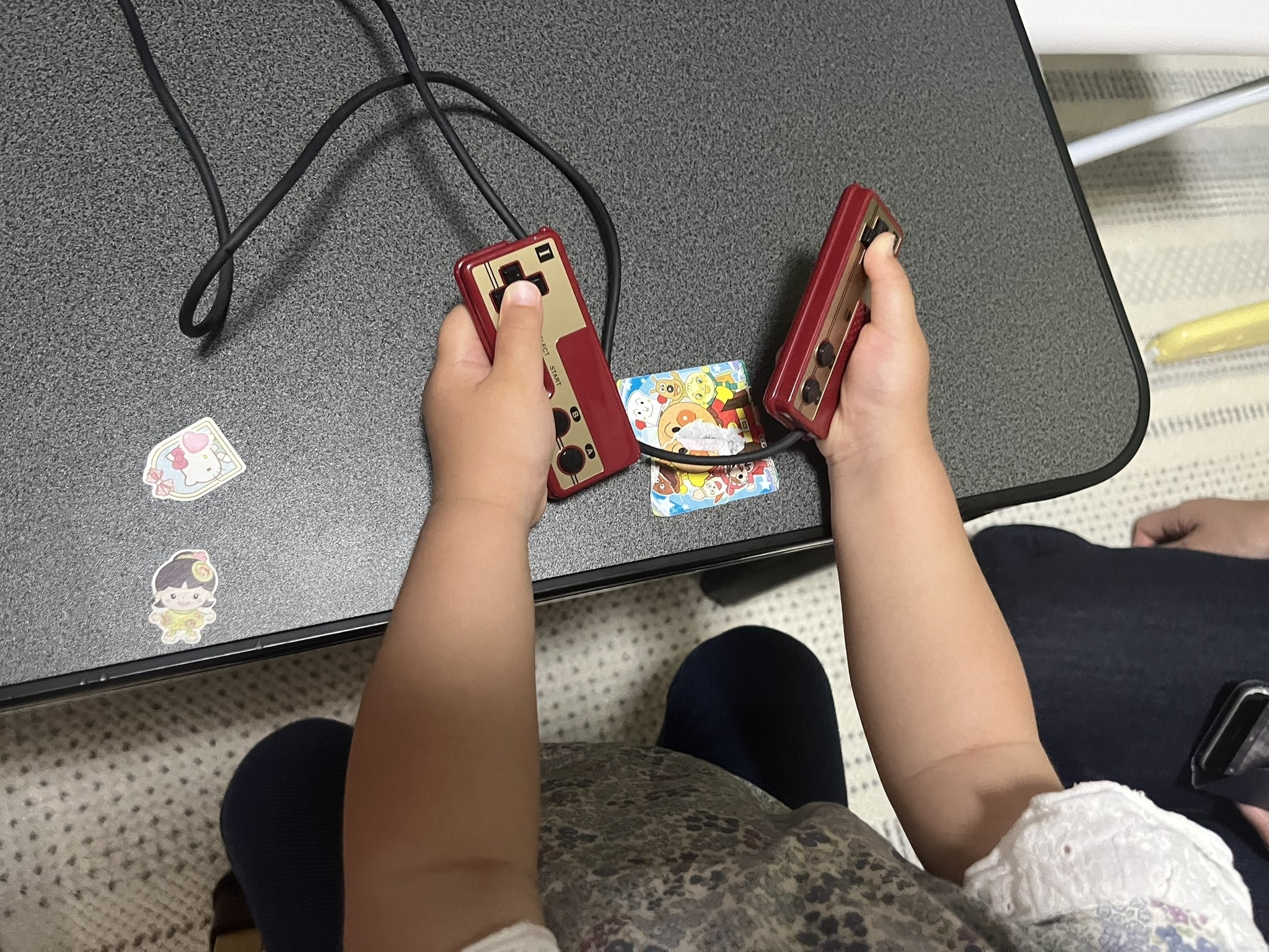 幼児がテーブルで2つのゲームコントローラーを持っています。テーブル上にはステッカーも数枚あります。