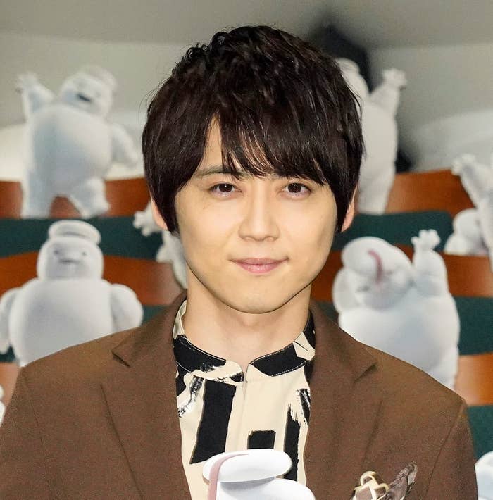 男性が後方のキャラクターたちと一緒にカメラに向かって微笑んでいます。彼はフォーマルなジャケットを着ています。